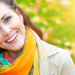 A woman with vibrant blue eyes and a beaming smile is wearing a colorful scarf and a light coat, hinting at autumnal weather. The background is a blur of golden autumn leaves and sunlight, giving the image a warm, joyful feel that matches her cheerful expression.