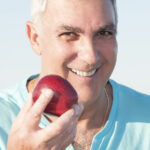 A smiling older man with gray hair is holding a red apple up to his mouth, preparing to take a bite. He's wearing a light blue shirt, and the bright background suggests a sunny, outdoor setting. The image conveys themes of healthy eating, wellness, and vitality in later life.