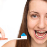 A cheerful young girl with braces is holding a toothbrush with blue toothpaste on it, ready to brush her teeth. Her sparkling eyes and open-mouthed smile showcase the metal braces on her teeth, emphasizing the importance of oral hygiene, especially during orthodontic treatment. The white background enhances her clean and healthy dental care routine.
