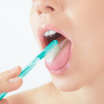 Close-up of a person brushing their teeth, showing a turquoise toothbrush with white bristles contacting the lower teeth. The person has glossy pink lips, and the image captures a moment of daily dental care with a focus on oral hygiene. The white background accentuates the clean and fresh atmosphere.