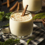 Homemade eggnog in a glass garnished with a cinnamon stick and a dusting of ground spice, typically nutmeg, is set on a rustic wooden table with additional cinnamon sticks and a festive evergreen branch nearby. A cozy holiday ambiance is evoked by the warm lighting and the classic checkered cloth beneath the glass.