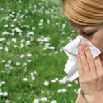 A woman with her blonde hair pulled back is using a tissue to blow her nose. She appears to be outdoors, with a blurry background of a green field dotted with white flowers, possibly indicating symptoms of hay fever or allergies. The focus is on her action and the potential discomfort she might be feeling.