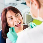 A young woman is sitting in a dental chair with her mouth open as a dentist prepares to administer an injection. The dentist, wearing a mask and green gloves, holds a syringe in one hand and a dental tool in the other. The woman appears calm and is looking at the dentist. The background shows a clean, modern dental office with cabinets and equipment. The image highlights a dental procedure, focusing on patient care and professionalism.