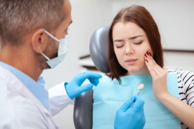 A woman sitting in a dental chair is touching her cheek with a pained expression, indicating dental discomfort. A dentist wearing a mask and blue gloves holds a dental mirror and speaks to her. The woman is wearing a striped shirt and a blue dental bib. The background shows a clinical setting, emphasizing a visit to the dentist for tooth pain or a dental check-up.