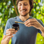 A smiling man with a beard is holding a bamboo toothbrush and a jar of charcoal toothpaste in a lush, green garden. He seems to be preparing to brush his teeth, illustrating an eco-friendly dental hygiene routine. The natural background adds to the theme of sustainability.