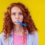 A young woman with curly red hair and wide eyes is brushing her teeth with a blue toothbrush, with toothpaste foam visible on her mouth. She's wearing a lavender shirt over a pink top, against a vivid yellow background, creating a playful and vibrant image that captures a moment of daily dental care with a humorous expression.