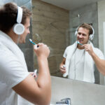 A young man with headphones on smiles while brushing his teeth, looking at his reflection in a modern bathroom mirror. He wears a white bathrobe and has a white towel draped over his shoulders. The bathroom has grey tiled walls and a glass shower enclosure. The image conveys a sense of casual, everyday grooming while enjoying music.
