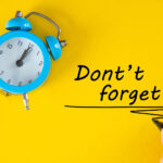 A classic blue alarm clock is positioned on the left side of a bright yellow background, showing a time of 10:09. Next to it, a hand with a black pen writes the phrase "Don't forget" as a reminder, combining the concepts of time management and memory.