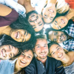 A group of eight diverse friends huddled in a circle, looking down towards the camera with bright smiles. The sunlight backlights the scene, highlighting their happy expressions and the bond of friendship.