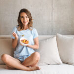 A smiling woman in a blue t-shirt sits cross-legged on a white couch, holding a bowl of fruit salad and a fork, with a concrete wall in the background. She embodies a relaxed and healthy lifestyle in a modern home setting.
