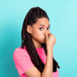 A woman with braided hair, wearing a pink shirt, playfully holding her nose, against a turquoise background, possibly reacting to a bad smell.