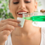Close-up of a woman smiling as she pours green mouthwash into a cap, preparing for her oral hygiene routine, with blurred greenery in the background.