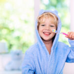 A cheerful young child with blond hair, wearing a blue hooded robe with ears, brushes their teeth with a pink toothbrush, with a bright, blurred background suggesting a sunny bathroom or bedroom.
