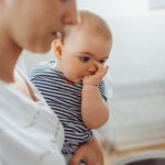 Close-up of a baby, held by a mother, sucking on their thumb while wearing a striped outfit, with a blurred kitchen sink in the background.