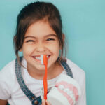 A cheerful young girl squinting happily with a toothy smile, holding a large toothbrush against a set of oversized model teeth. She's wearing a white shirt with a blue and brown jumper, emphasizing dental hygiene in a playful manner against a soft teal background.