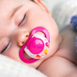 A peaceful sleeping baby with a pink pacifier in their mouth, nestled in soft white bedding. The baby's delicate eyelashes are highlighted in a close-up, and a hint of a cozy blue and purple knit blanket adds a touch of warmth to the tranquil scene.