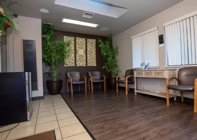 A waiting room with a warm and welcoming atmosphere, featuring a wooden floor, several brown leather chairs with wooden arms, and a reception desk. The room is adorned with potted bamboo plants and a decorative wall panel. Sunlight filters through vertical blinds, contributing to the serene and comfortable setting.