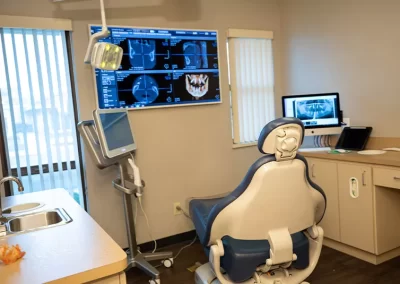 A well-equipped dental examination room with an empty patient chair, dental light, and a sink. On the wall, a large screen displays detailed dental CT scans, while a secondary monitor shows additional X-ray images. The room is designed for comfort and efficiency, with natural light coming from a window with blinds.