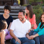 A joyful family moment with a man seated on steps surrounded by four young women, possibly his daughters. They are all smiling and engaging in a relaxed outdoor setting, suggesting a strong family bond and a happy gathering.