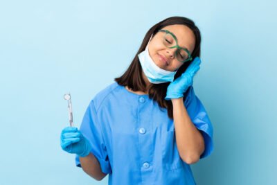 A tired female dentist in a blue scrub, wearing glasses and a surgical mask pulled down to her chin, is holding a dental mirror and rubbing her neck with her other hand, indicating fatigue or stress. The solid light blue background enhances her expression of exhaustion.