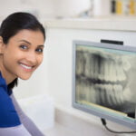 A smiling dental professional in a navy blue uniform sitting beside a computer monitor displaying dental X-rays, suggesting a positive and professional dental consultation environment.
