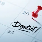 White calendar with a red push pin on the 20th with the word Dentist! written in for an appointment.