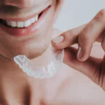 A close up image of a man smiling while holding a mouthguard in the left hand. Mouthguards help protect your teeth against teeth grinding as well as during sports.