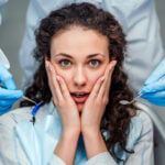 A young woman with curly hair sits in a dentist's chair, looking startled and holding her face with both hands. A dentist, whose face is not visible, stands behind her, wearing a white lab coat and blue gloves, holding dental tools close to her face. The woman wears a dental bib, and her expression suggests anxiety or fear.