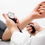 A healthcare professional checks a patient's blood pressure using a sphygmomanometer. The patient has their arm extended with a cuff wrapped around it, while the healthcare provider holds the pressure gauge and bulb, monitoring the reading. The background is plain and white, focusing attention on the procedure.