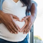 A close-up of a pregnant woman with her partner standing behind her, both forming a heart shape over her belly with their hands. The woman is wearing a white shirt and blue cardigan, and the scene conveys warmth, support, and anticipation.