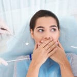 A woman sits in a dental chair, covering her mouth with both hands and looking anxious. Three gloved hands holding dental instruments, including a mirror and probe, surround her, emphasizing her nervousness about the dental visit. She is wearing a blue protective bib.