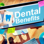 A computer monitor on a wooden desk displays a "Dental Benefits" advertisement featuring a doctor holding a clipboard with a large blue tooth graphic. The desk includes a keyboard, mouse, coffee cup, ruler, colorful paper clips, a rubber band ball, folders, eyeglasses, and sticky notes reading "Dr. Appt 2:00 p.m.", "Plan Life", and "Research - Marketing". A person's hands are visible using the keyboard and mouse. A green plant is in the upper left corner.