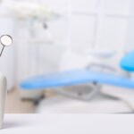 A white tooth model holding a dental mirror and scaler in the foreground, with a blurred blue dental chair in the background.